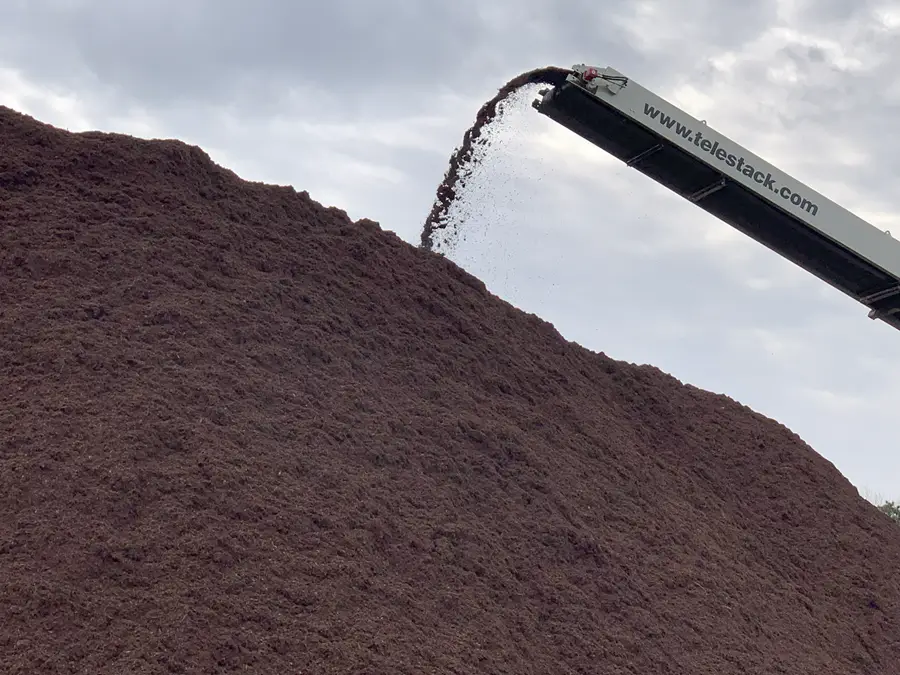Machinery unloading a large pile of soil or mulch under a cloudy sky.